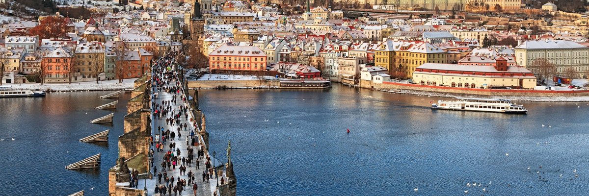 People walking across a bridge in Prague with the city and castle on the hill in the background.