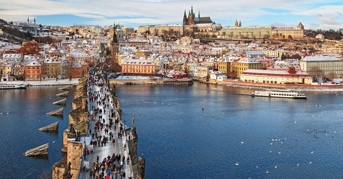 People walking across a bridge in Prague with the city and castle on the hill in the background.