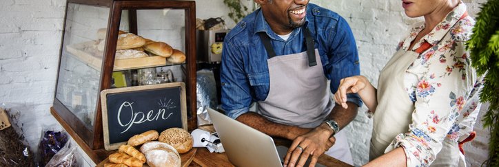 A cafe owner standing behind the counter with a laptop and joking with his employee.