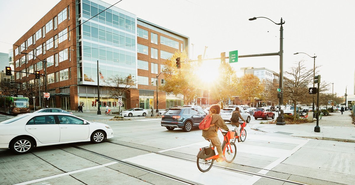 Cars driving through stoplights on a busy street as bikes ride by.