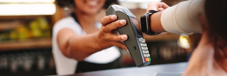 A smiling cashier holding up a payment reader for a woman paying with her smart watch.