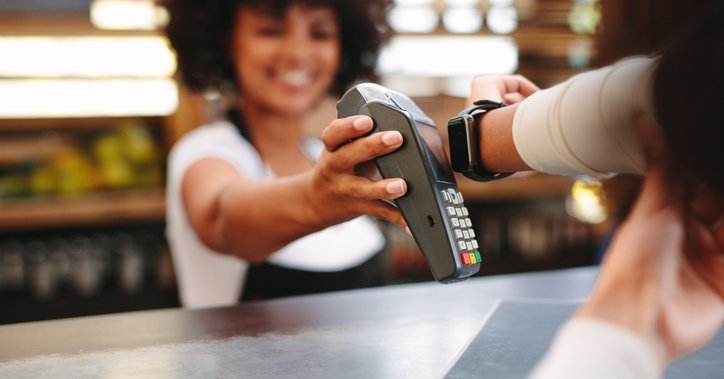 A smiling cashier holding up a payment reader for a woman paying with her smart watch.