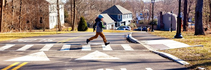 A child running across a suburban street with houses in the background and the word SLOW painted on the street in the foreground.