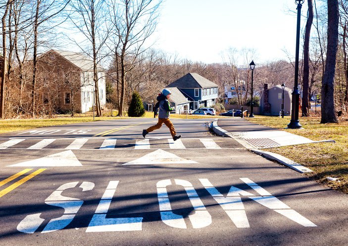 A child running across a suburban street with houses in the background and the word SLOW painted on the street in the foreground.