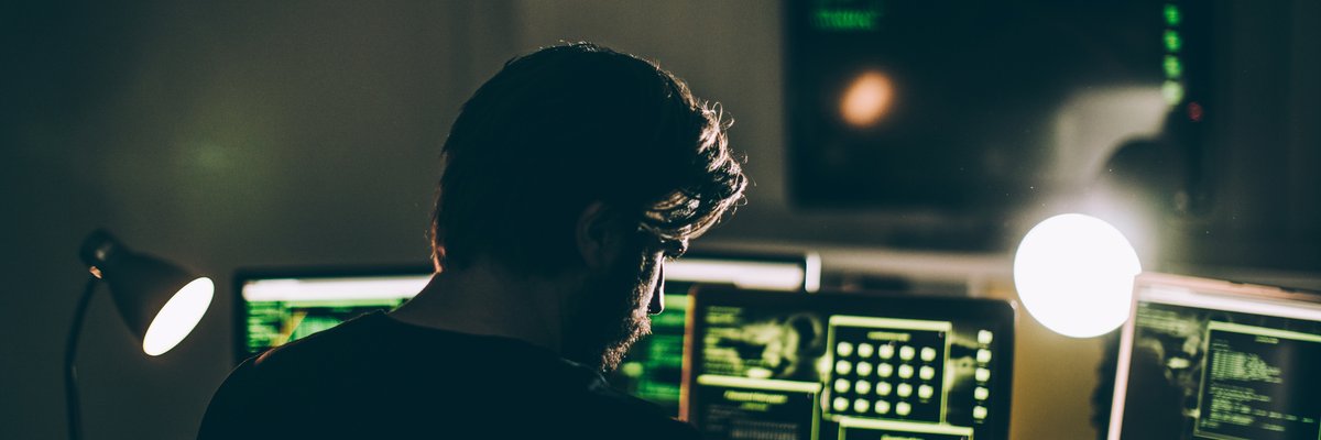 A computer hacker with multiple monitors in a dark room.