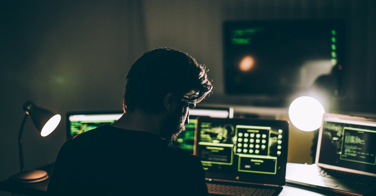 A computer hacker with multiple monitors in a dark room.