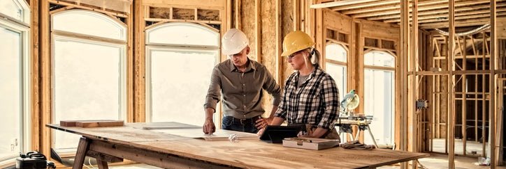 Two construction workers looking at plans on a table in a house being built.