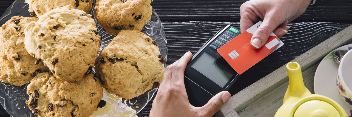 A bakery customer swiping a credit card next to a plate of muffins and a teapot.
