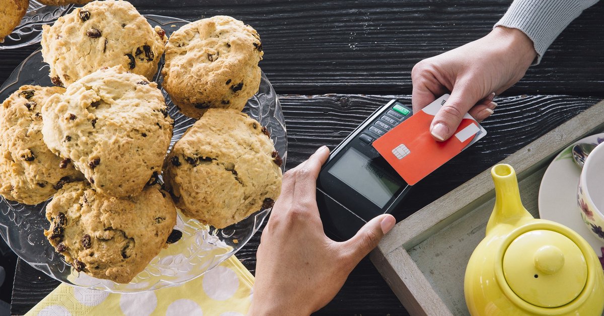 A bakery customer swiping a credit card next to a plate of muffins and a teapot.