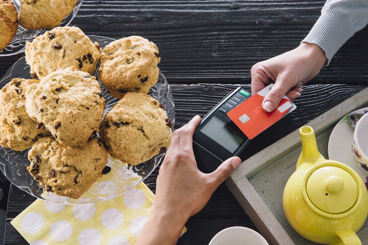 A bakery customer swiping a credit card next to a plate of muffins and a teapot.