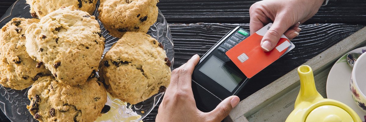 A bakery customer swiping a credit card next to a plate of muffins and a teapot.