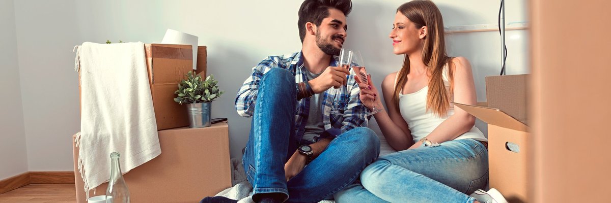 young couple sitting on floor of empty apartment surrounded by boxes and toasting with champagne