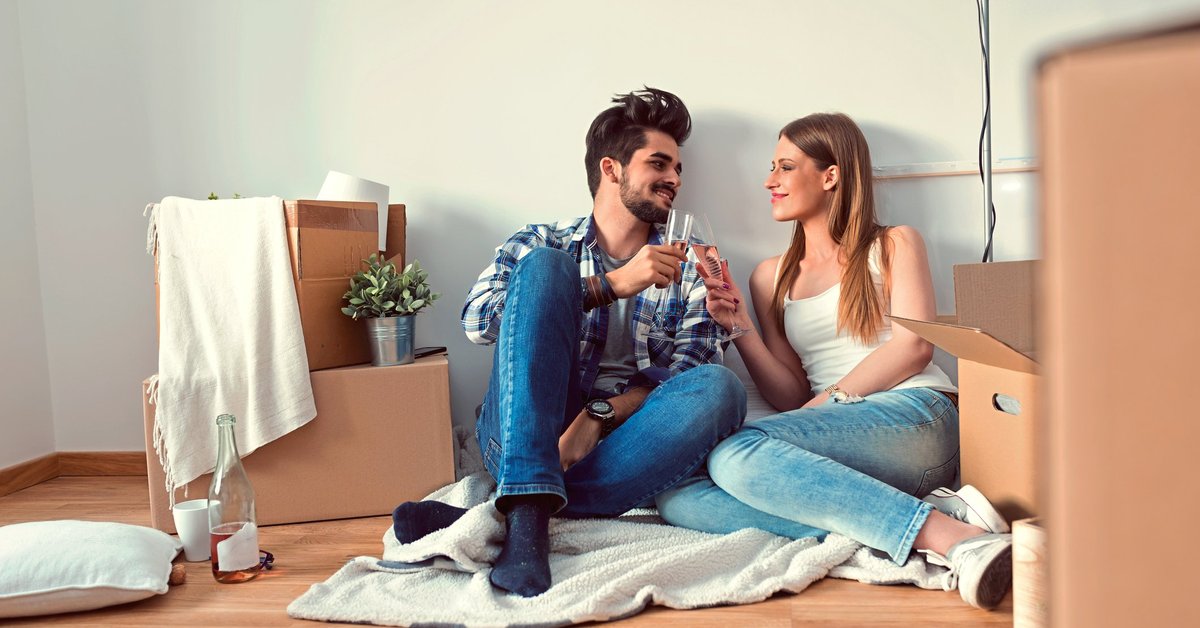 young couple sitting on floor of empty apartment surrounded by boxes and toasting with champagne
