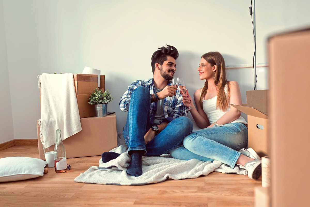young couple sitting on floor of empty apartment surrounded by boxes and toasting with champagne