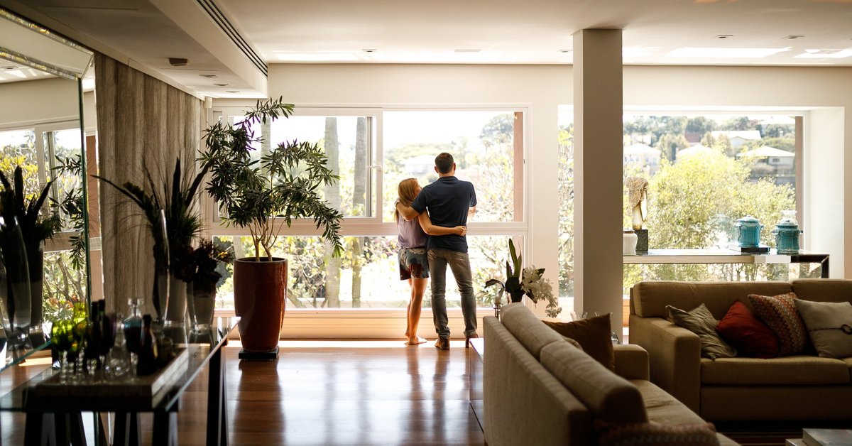 A couple with their arms around each other at the window of their home.