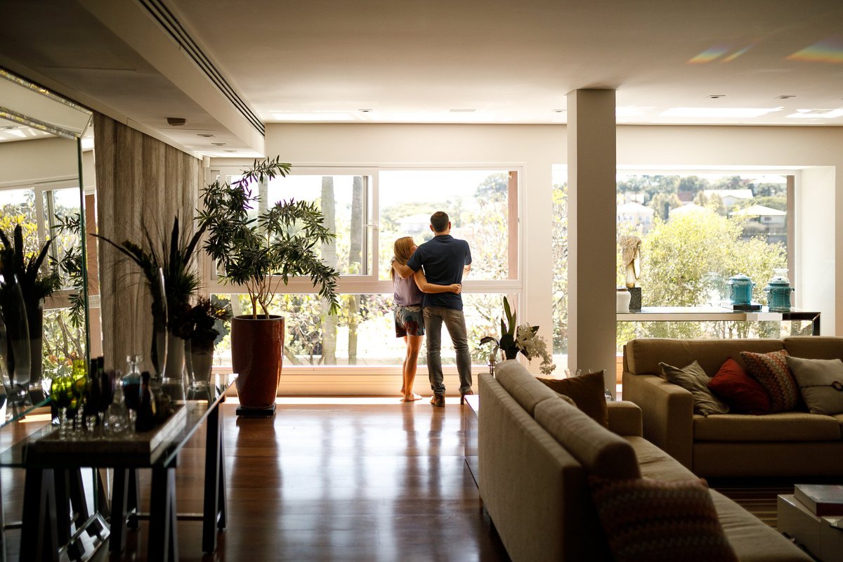 A couple with their arms around each other at the window of their home.