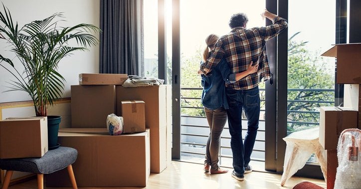 A man and woman standing with their arms around each other looking out a large window with moving boxes in the living room behind them.
