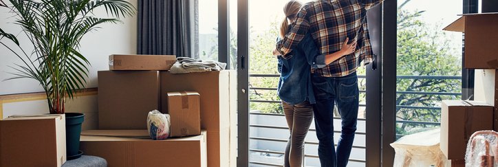 A man and woman standing with their arms around each other looking out a large window with moving boxes in the living room behind them.