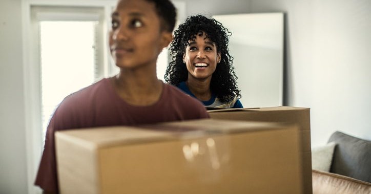 Two women looking around while carrying moving boxes into their new home.
