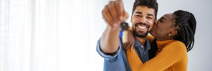 A woman kissing a smiling man's cheek while he holds up keys to the new house they're standing in.