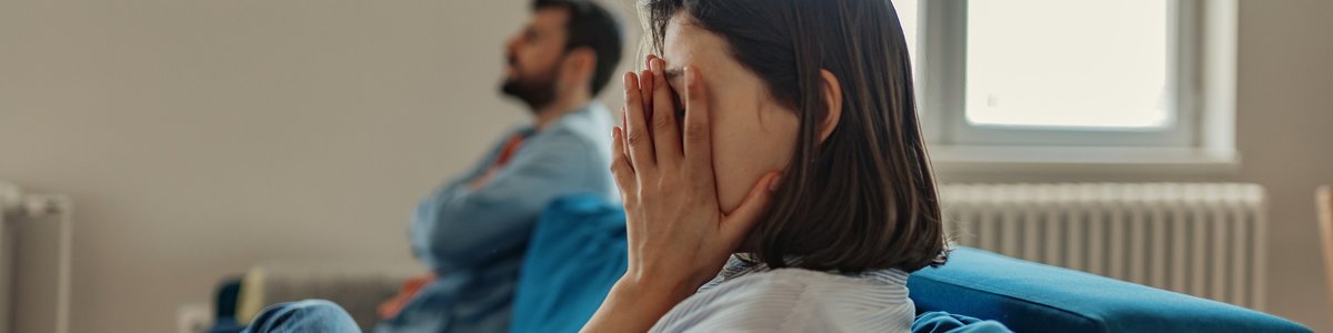 A woman sitting at one end of a couch with her hands covering her face while a man sits with his arms crossed on the other end.