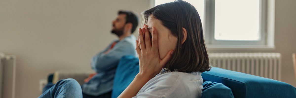 A woman sitting at one end of a couch with her hands covering her face while a man sits with his arms crossed on the other end.