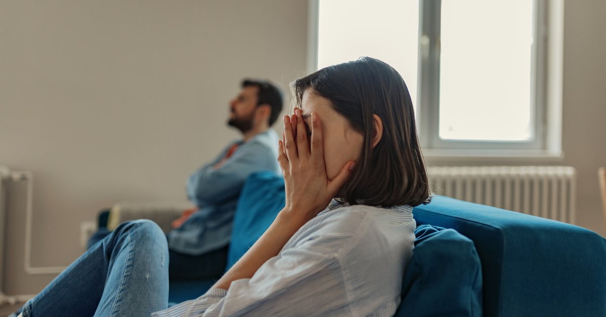 A woman sitting at one end of a couch with her hands covering her face while a man sits with his arms crossed on the other end.
