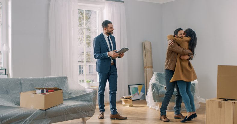 A man and woman hugging in the living room of their new house surrounded by moving boxes standing next to their realtor.