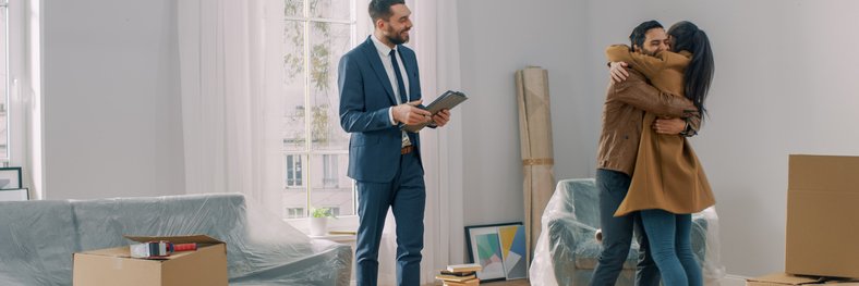 A man and woman hugging in the living room of their new house surrounded by moving boxes standing next to their realtor.