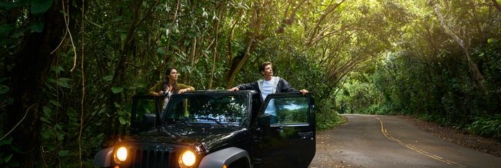 A man and woman standing in the open doors of their car parked on the side of a forest road.