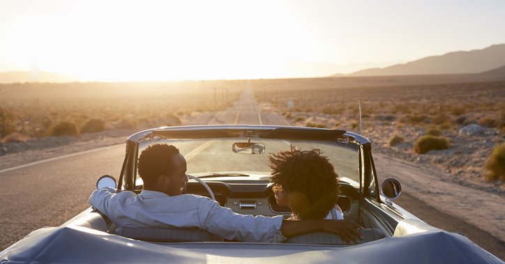 A smiling couple driving their car down an open highway in the desert at dusk.