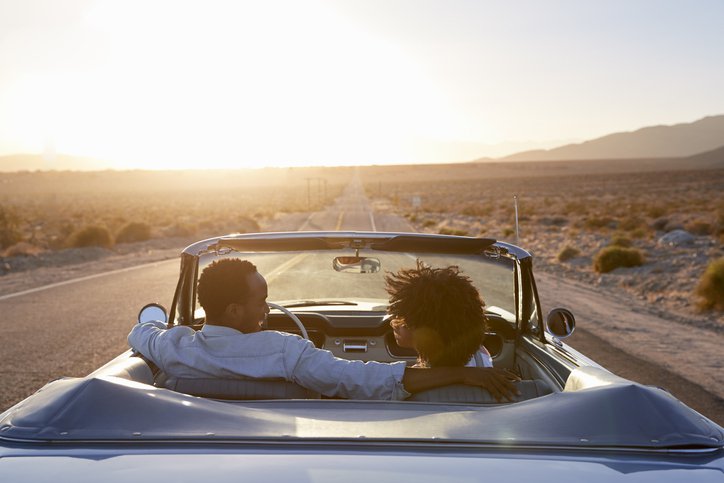 A smiling couple driving their car down an open highway in the desert at dusk.