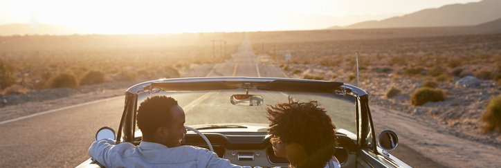 A smiling couple driving their car down an open highway in the desert at dusk.