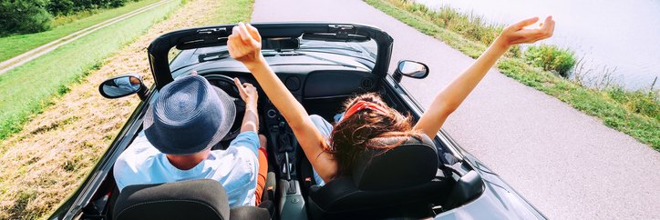 A man and woman driving down a scenic road next to a lake with the convertible top down.