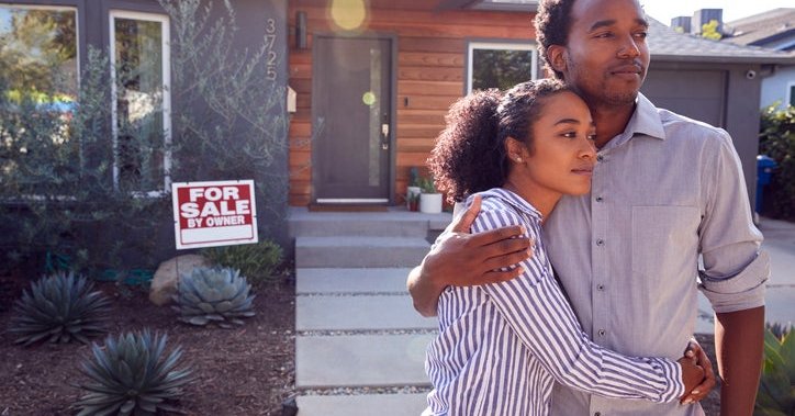 A man and woman standing with their arms around each other in front of their house with a For Sale sign behind them.
