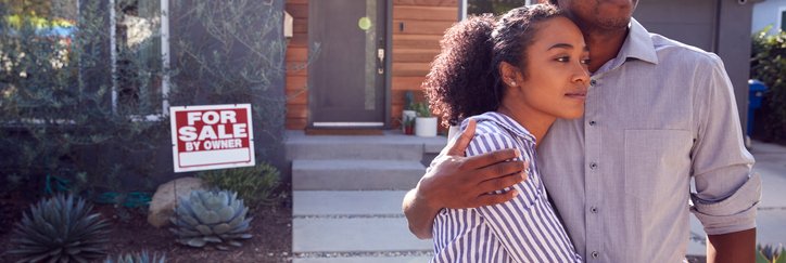 A man and woman standing with their arms around each other in front of their house with a For Sale sign behind them.