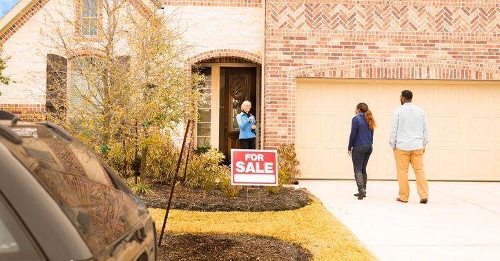 A man and woman meeting their realtor at the front door of a house with a For Sale sign out front.