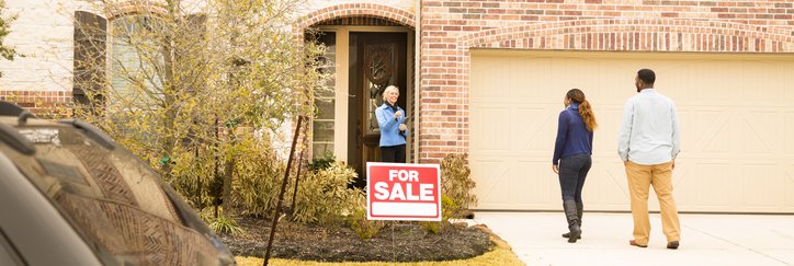 A man and woman meeting their realtor at the front door of a house with a For Sale sign out front.
