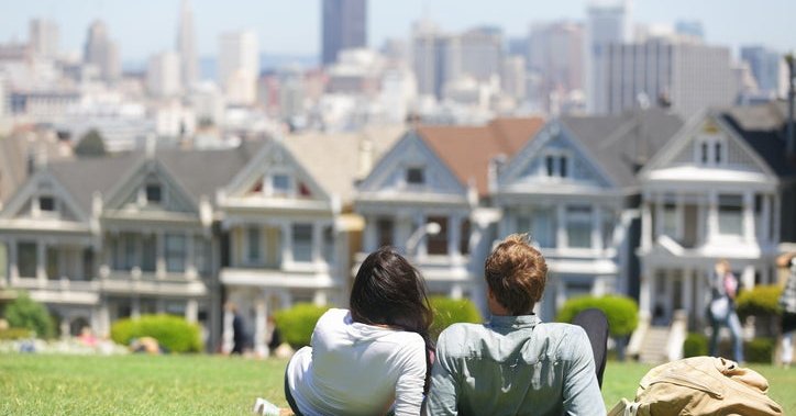 A man and woman lying in the grass in a park on a sunny day facing the Painted Ladies houses in San Francisco.
