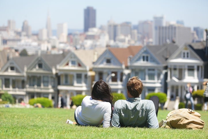 A man and woman lying in the grass in a park on a sunny day facing the Painted Ladies houses in San Francisco.