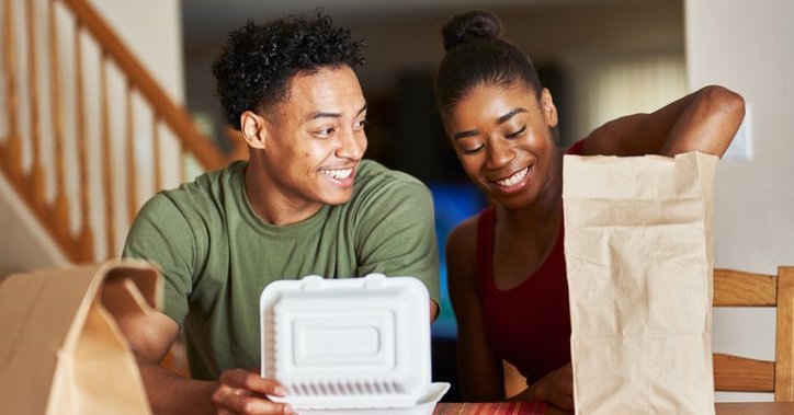 A man and woman sitting at their dining table and opening takeout food containers.
