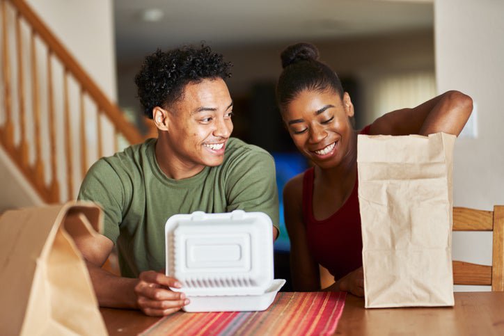 A man and woman sitting at their dining table and opening takeout food containers.