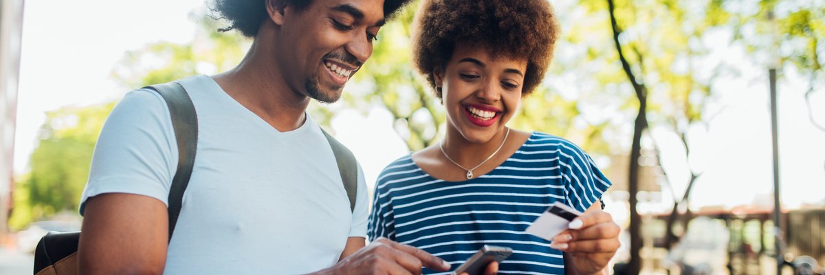 A smiling couple standing outside and holding a phone and credit card.