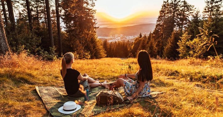 Two women sitting on a picnic blanket on a tree-lined hill overlooking a sunset.