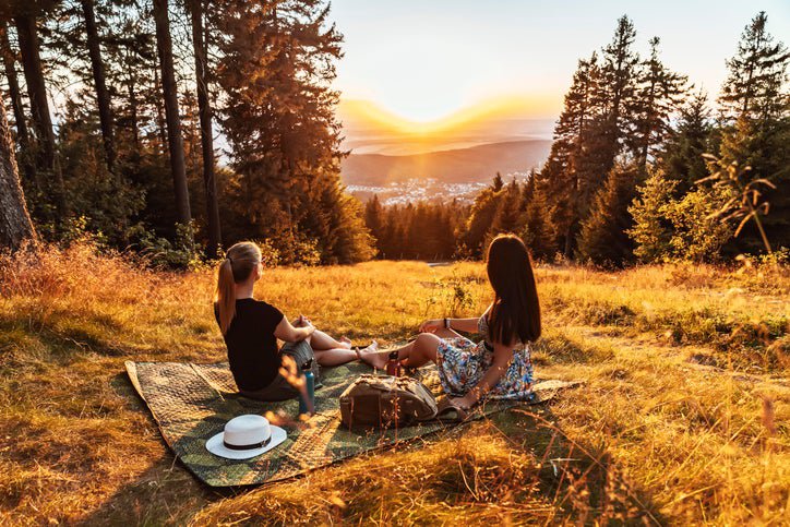 Two women sitting on a picnic blanket on a wooded hill overlooking a sunset.