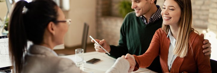 A man and woman signing financial documents while sitting at a desk and shaking hands with a business professional.