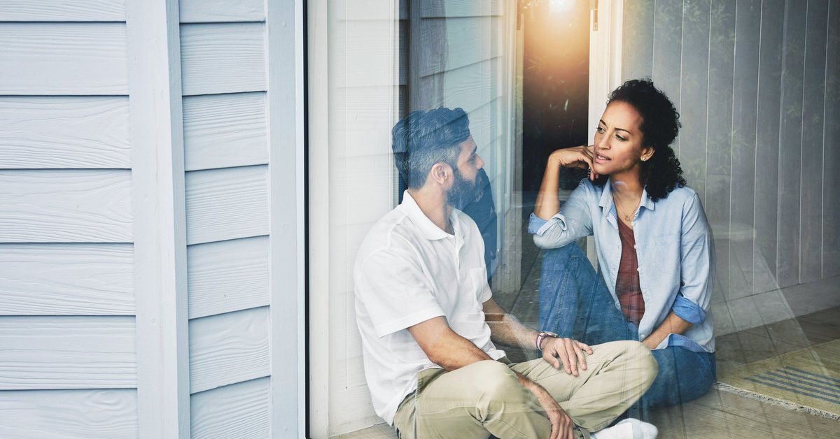 A young couple having a conversation while sitting on the floor in front of a sunny window.
