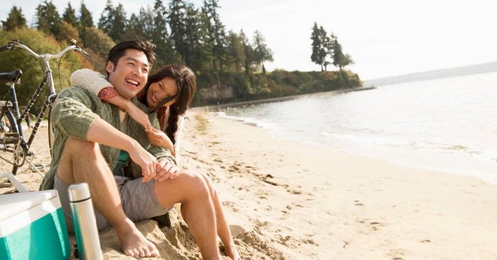 A smiling man and woman sitting next to a bike and ice chest at the beach with their arms around each other.