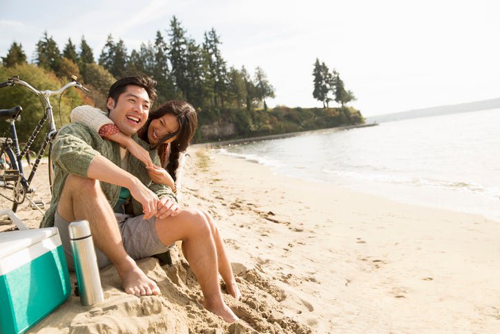 A smiling man and woman sitting next to a bike and ice chest at the beach with their arms around each other.