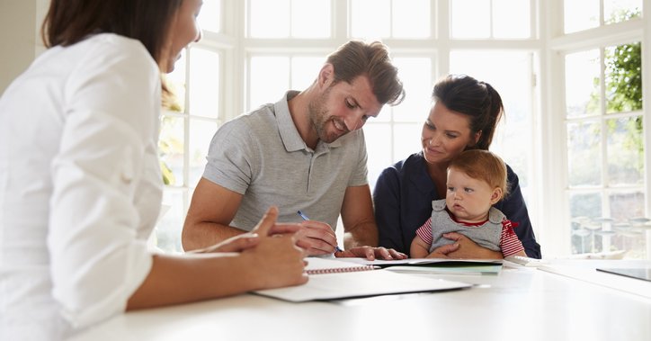 A man sitting next to his wife and baby at a sunny kitchen counter and signing documents from a female mortgage lender.